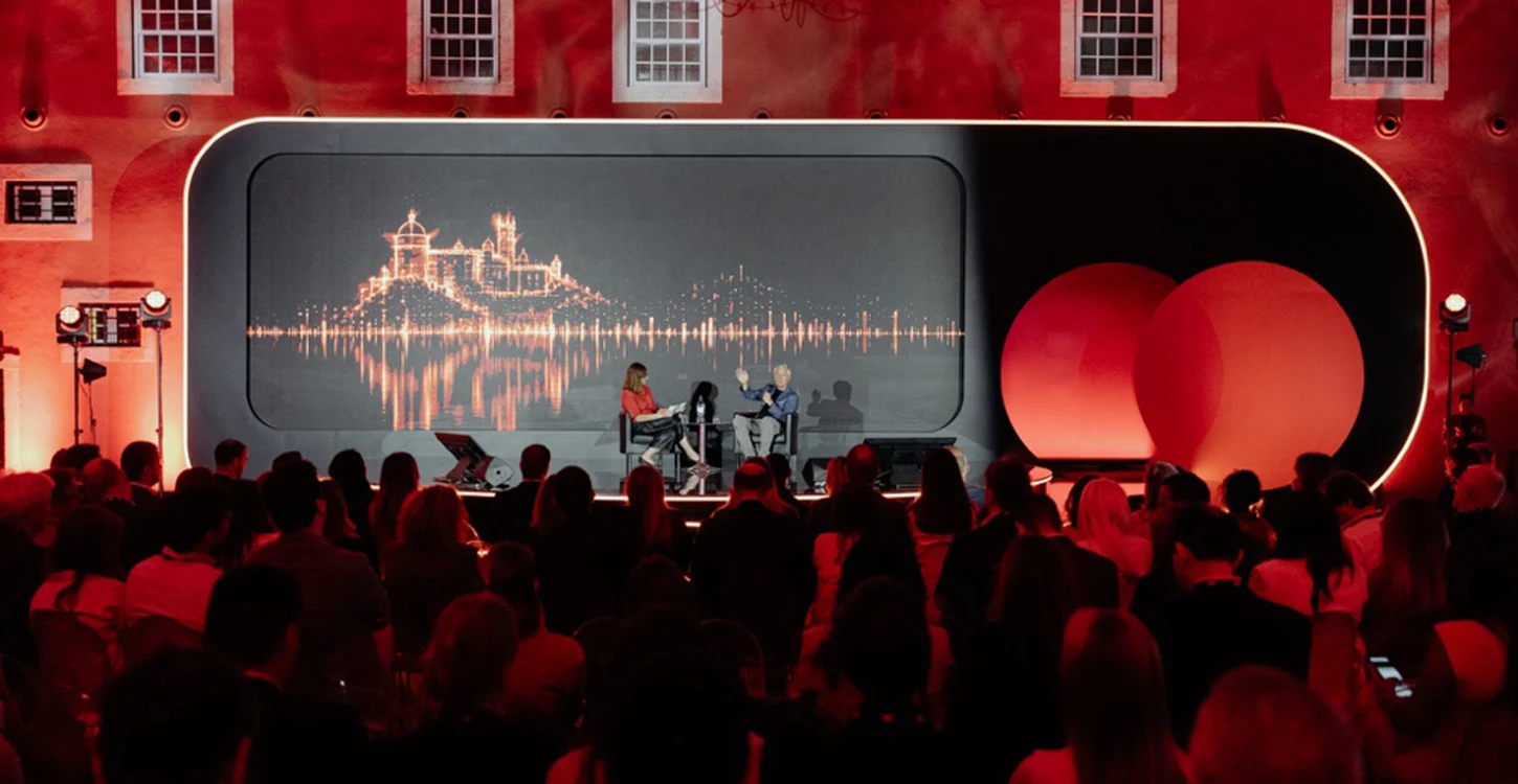 Palestra num palco diante de uma plateia, com projeção de um castelo e formas de onda sonoras ao fundo