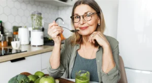 Mulher numa cozinha a provar uma colher de sopa, junto a fruta, brócolos e um batido verde