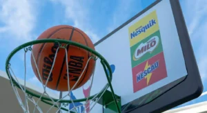Bola de basquetebol no cesto, diante de uma tabela com logótipos Nesquik, Milo e Nescau