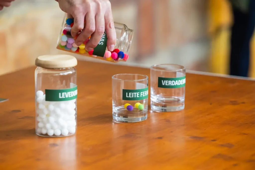 Mão despeja bolas coloridas de um frasco para copos rotulados com “LEVEDURA”, “LEITE FERMENTADO” e “VERDADE”