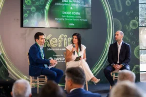 Painel com três participantes num evento, com uma mulher a falar ao microfone perante uma audiência