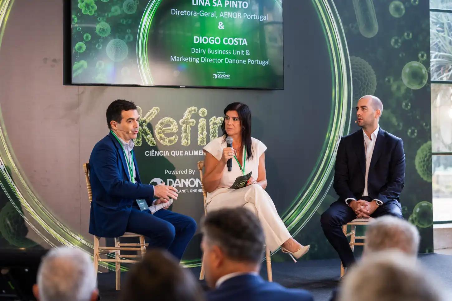 Painel com três participantes num evento, com uma mulher a falar ao microfone perante uma audiência