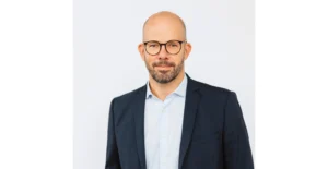 Homem careca, de óculos e barba, vestido com blazer escuro e camisa clara