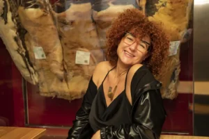 Mulher de cabelo encaracolado e óculos, sorrindo diante de grandes peças expostas numa vitrina