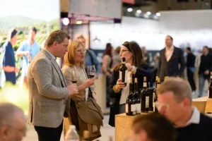 Três pessoas conversam numa prova de vinhos, enquanto uma mulher apresenta uma garrafa a um casal