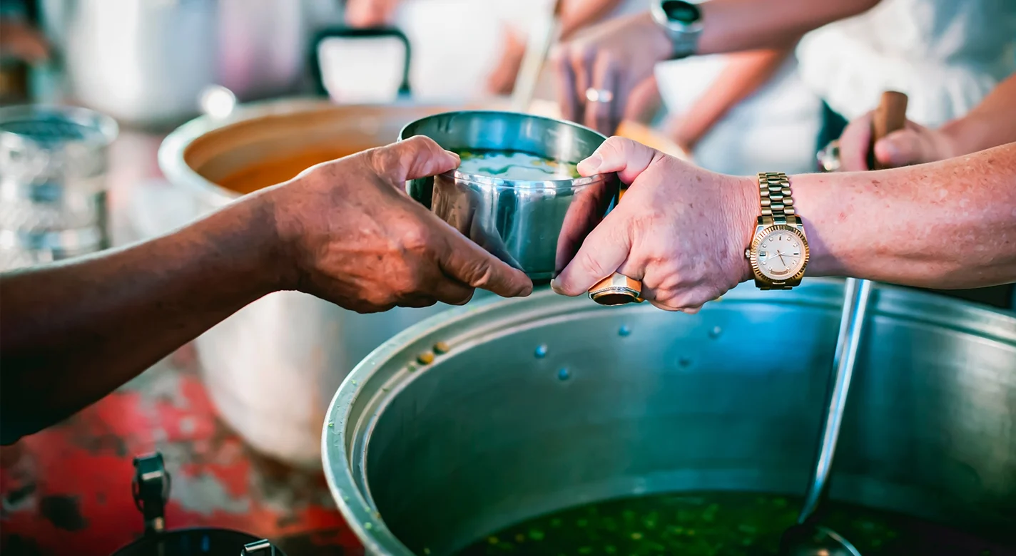 Duas pessoas passam uma taça de metal sobre um grande recipiente com sopa