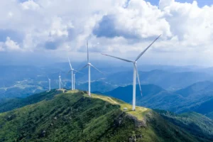 Aerogeradores num cume verdejante, com montanhas e nuvens ao fundo